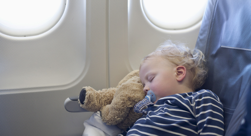 Bebé dormido con chupete y peluche en un asiento de avión, durante un vuelo diurno, transmitiendo tranquilidad y confort en un viaje familiar.
