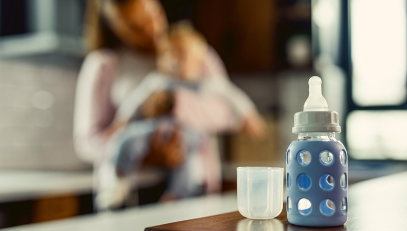 Biberón preparado con leche de fórmula en una cocina, con una madre sosteniendo a su bebé al fondo