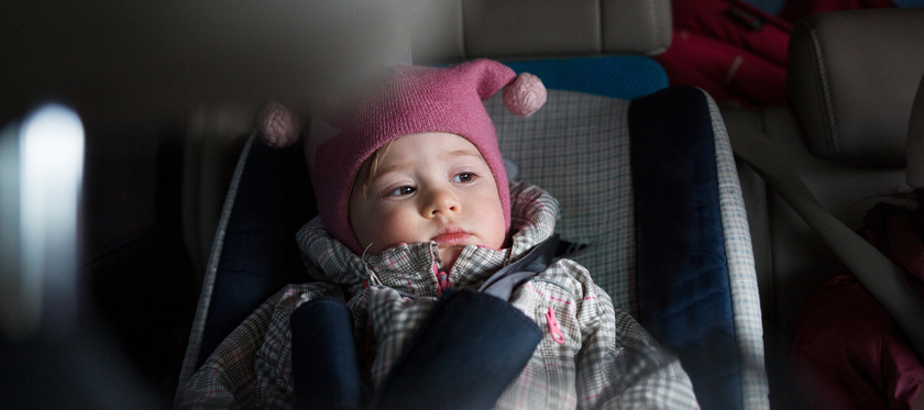 Niña pequeña con gorro rosa sentada en su silla de coche, con expresión seria, abrochada con el cinturón de seguridad durante un viaje en coche.